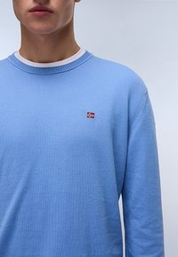 Suéter azul claro con un cuello redondo, textura de punto y un pequeño logo de bandera bordado en rojo y blanco en el pecho. Mangas largas visibles.