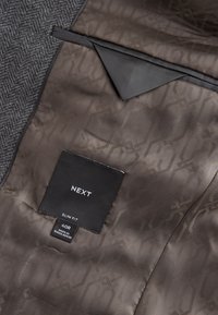 Doublure de veste marron à motif avec une étiquette noire indiquant « NEXT Slim Fit 40R Made in Bangladesh » et bord en tissu chevrons gris.