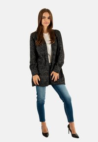 Ung kvinde står selvsikkert iført en sort strikket cardigan, hvid skjorte, blå jeans og sorte høje hæle foran en ensfarvet baggrund.
