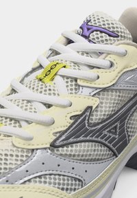 Zbliżenie białego i szarego buta sportowego z siateczkową tkaniną, żółtymi akcentami, białymi sznurowadłami oraz czarnym logo na boku.