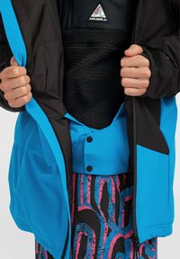Chaqueta aislante azul y negra con forro de polar. Presenta un cierre con cremallera, botones a presión y una capa interior negra texturizada.