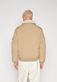 Una chaqueta bomber beige con un cuello texturizado y esponjoso. Presenta puños y cinturilla elásticos, con una tela exterior suave y un diseño minimalista.