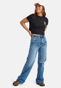 Zwarte cropped t-shirt met een graphic op de linkerborst, gecombineerd met lichtblauwe, losse hoge taille jeans en bruine schoenen.