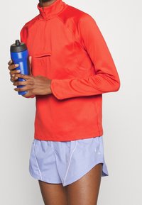 Rotes sportliches Quarter-Zip-Oberteil mit hohem Kragen, kombiniert mit hellvioletten Shorts mit weißen Akzenten. Wasserflasche in der Hand.