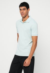 Világoskék póló puha anyagból, gallérral, rövid ujjakkal és egy finom logóval. Sötét farmerrel viselve.