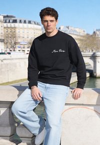 Jeune homme en sweat-shirt noir « Mira Paris » et jean clair appuyé sur la rambarde en pierre d’un pont parisien avec une rivière et des bâtiments en arrière-plan.