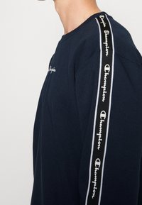 Marineblauwe crewneck sweatshirt met het witte Champion-logo op de borst en langs de zijkant, met een zachte katoenen textuur en een reguliere pasvorm.