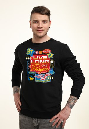 Mężczyzna w czarnej bluzie z kolorowym wzorem Star Trek i napisem „Live Long and Prosper”, stojący z rękami na biodrach.