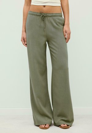 Personne portant un pantalon large vert olive avec une taille à cordon et des sandales beige, debout devant un fond uni clair.