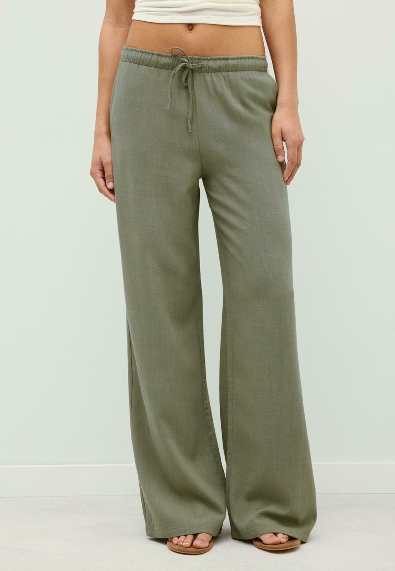 Personne portant un pantalon large vert olive avec une taille à cordon et des sandales beige, debout devant un fond uni clair.
