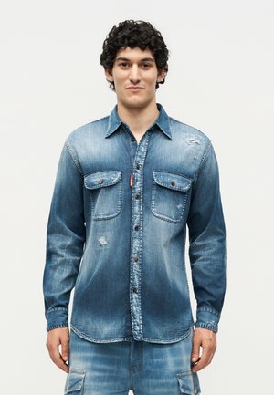 Νέος άνδρας με σγουρά μαλλιά, φορώντας ένα ξεθωριασμένο μπλε denim πουκάμισο με τσέπες στο στήθος και ταιριαστό denim παντελόνι cargo, στέκεται μπροστά σε απλό φόντο.