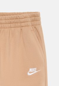 Beige Nike-housut, joissa on joustava vyötärö, sivutaskut ja valkoinen brodeerattu logo vasemmalla alhaalla. Pehmeä ja sileä rakenne.