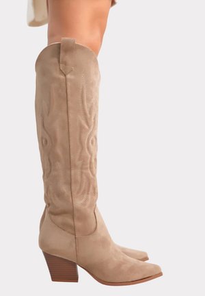 Bottes de cowboy hautes en daim beige avec motifs décoratifs cousus, talons empilés en bois, portées sur des jambes nues sur un fond neutre.