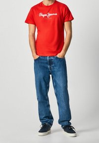 Camiseta de algodón roja con logo blanco de "Pepe Jeans London", combinada con unos vaqueros de mezclilla azul y zapatillas negras. Ajuste casual y diseño sencillo.