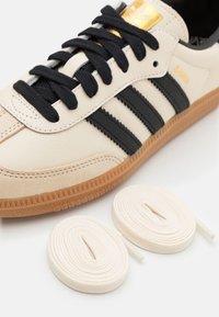 Cream leather sneaker with black stripes, textured gum sole, and gold logo detailing. Includes two pairs of white laces.