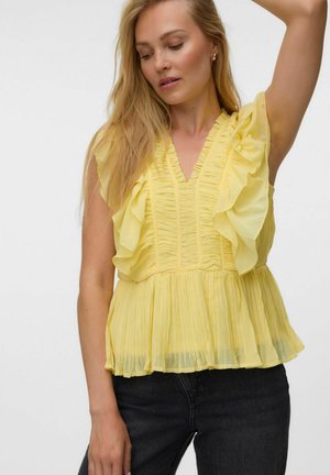 Femme portant un chemisier jaune sans manches à volants avec des plis, debout, un bras levé, regardant vers le bas.