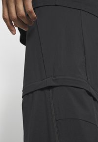 Zwarte broek met een slim fit en een gladde textuur, met zichtbare stiksels, een schuine zak en een ritsdetail aan de zijkant.