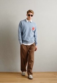 Light blue sweatshirt with a circular red graphic, white collared shirt underneath, brown wide-leg trousers, and gray sneakers.