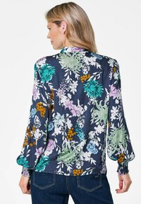 Blumige Bluse mit langen, puffigen Ärmeln. Hat einen navyfarbenen Hintergrund und ist mit grünen, lila und gelben Blumen sowie einem gestuften Manschettendesign versehen.