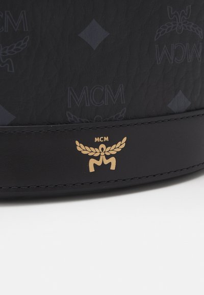 Sac en cuir noir avec un motif subtil du logo MCM, présentant un emblème doré et un détail de couture à la base. Texture lisse, design élégant.