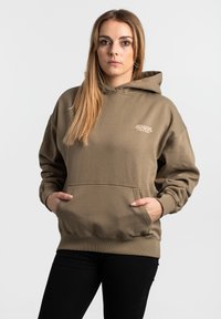 Hnedá mikina s kapucňou, predným vreckom, sťahovacou šnúrkou, rebrovanými manžetami a lemom, s bielym logom na ľavej hrudi. Mäkký, textúrovaný materiál.