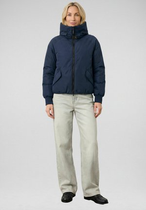 Vrouw in een marineblauwe donsjas met hoge kraag, lichtgrijze rechtvallende jeans en zwarte schoenen, staand tegen een effen achtergrond.