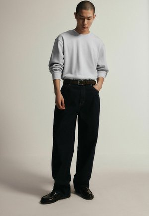 Man wearing light gray long-sleeve shirt, black wide-leg pants with belt, and black shoes, standing with one hand in pocket against plain background.