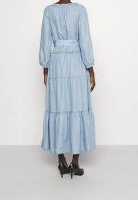 Ljusblå denim maxiklänning med långa puffärmar, elastisk midja, flera lager i kjolen och detaljerad midja. Bärs med svarta högklackade skor.