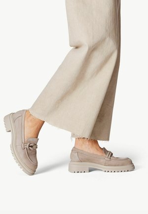 Personne portant un pantalon beige à jambes larges et des mocassins taupe à semelle épaisse avec un détail de nœud sur fond blanc.