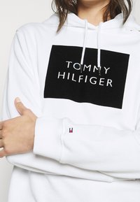 Felpa bianca con cappuccio che presenta un'etichetta rettangolare nera con "TOMMY HILFIGER" in grassetto bianco, cordini al collo, polsini a coste.