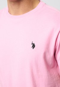 Lichtroze T-shirt met ronde hals en een klein zwart geborduurd logo van een polo speler te paard vlak bij de borst.