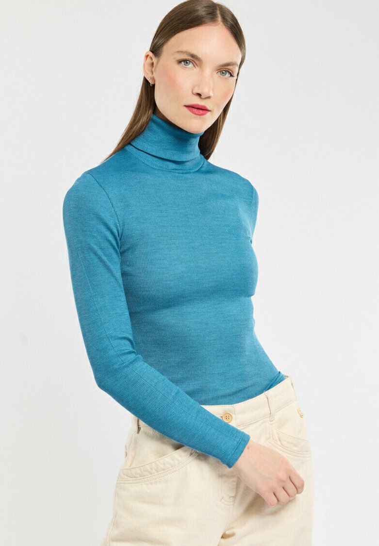 Türkiser Rollkragenpullover in Rippenstruktur, taillierte Form, mit langen Ärmeln, kombiniert mit hellbeigen, hochgeschnittenen Hosen mit Knöpfen.