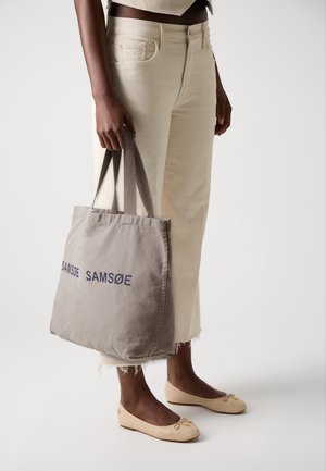 Gray canvas tote bag with "SAMSOE" printed in dark blue, held by a person wearing cream-colored jeans and beige flats.