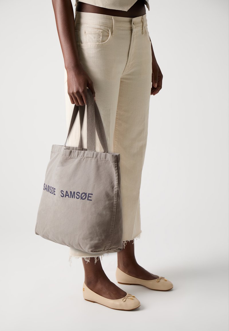Borsa tote grigia in canvas con la scritta "SAMSOE" stampata in blu scuro, tenuta da una persona che indossa jeans color crema e mocassini beige.