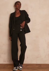 Blazer nero e pantaloni a vita alta con dettagli di bottoni lucidi, abbinati a scarpe sportive nere e bianche su un pavimento in legno.