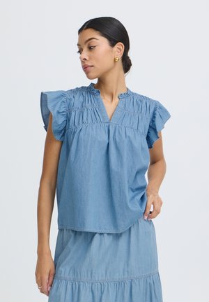 Žena oblečená v svetlomodrej blúzke s volánikovými rukávmi a rovnakej sukni, pozerá sa doľava, vlasy zviazané dozadu, zlaté náušnice.