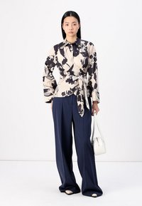 Blusa avvolgente in crema con motivo floreale nero, maniche lunghe e dettaglio legato in vita; abbinata a pantaloni navy a gamba larga e una borsa bianca.