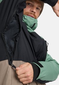 Multi-color jacket featuring a black upper, green sleeves, and a tan lower section. Includes a zipper, mesh lining, and Velcro cuff closures.