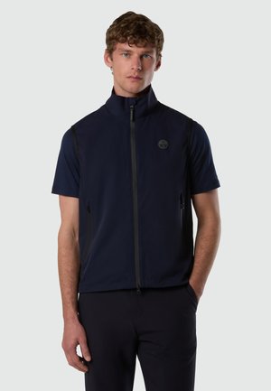 Giovane uomo con capelli corti e ricci che indossa un gilet blu navy senza maniche con cerniera sopra una maglietta abbinata a maniche corte, in piedi con una mano in tasca.