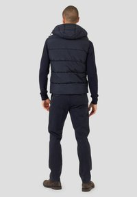 Chaleco acolchado azul marino con capucha, con paneles acolchados y bolsillos laterales con cremallera, combinado con pantalones oscuros y botas marrones. Diseño simple y práctico.