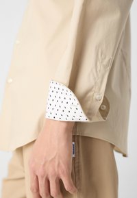 La manche d'une chemise beige montre un poignet blanc avec un petit motif géométrique noir, porté sur un pantalon beige assorti. La main est détendue sur le côté.