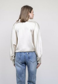 Chemisier satiné beige clair avec des manches bouffantes, associé à un jean en denim bleu légèrement délavé. Texture lisse et coupe ajustée visibles.