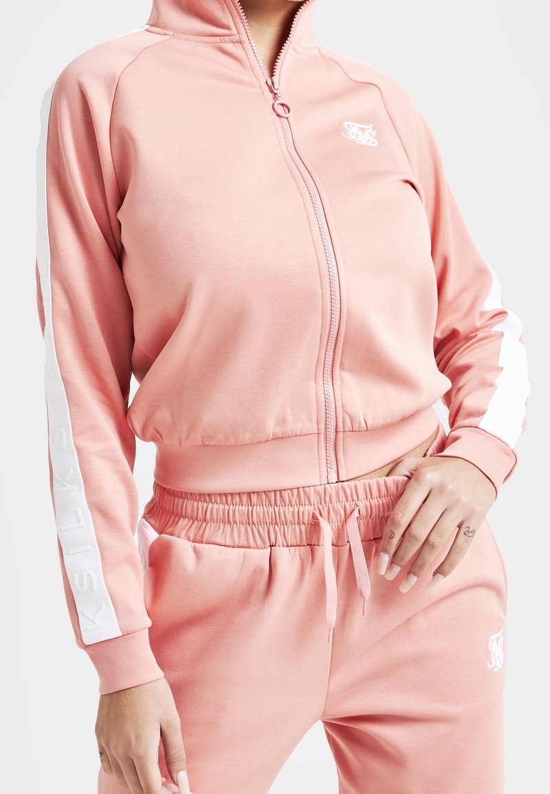 Femme portant une veste de survêtement zippée rose clair et un pantalon de survêtement assorti à taille élastique, avec logo blanc et détails de bandes sur les manches.