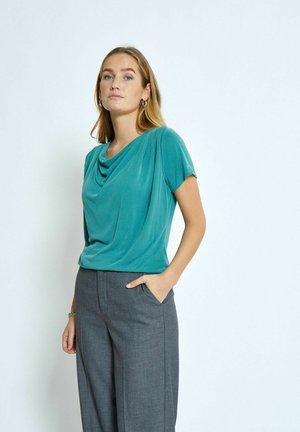 Top corto a maniche corte di colore teal con scollatura drappeggiata, abbinato a pantaloni grigi su misura. Tessuto liscio con una vestibilità rilassata e una leggera lucentezza.