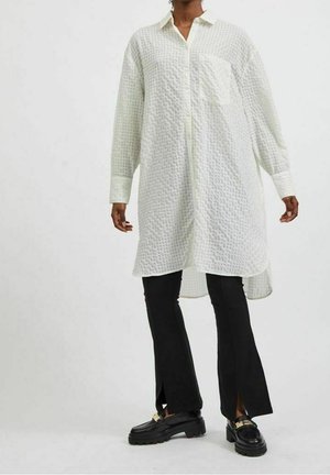 Persoon draagt een lang wit gestructureerd overhemd met knopen, zwarte uitlopende broek met voorste splitten en zwarte loafers met gouden beslag.