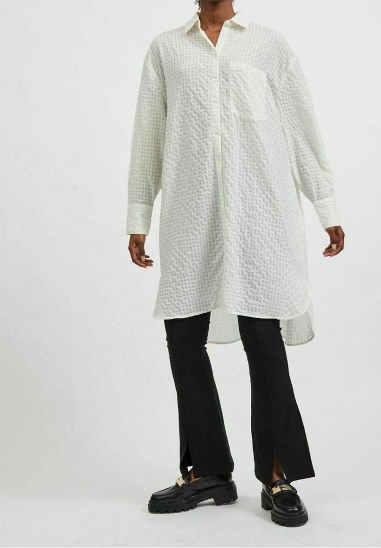 Personne portant une longue chemise blanche texturée à boutons, un pantalon noir évasé avec des fentes à l'avant, et des mocassins noirs ornés de ferrures dorées.