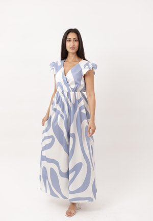 Femme portant une robe longue blanche et bleu clair à motifs avec des manches volantées, sur un fond blanc uni.