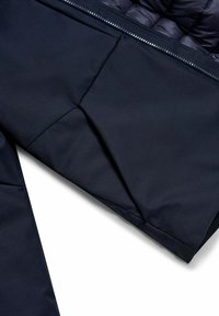 Primo piano di una manica di giacca blu scuro e zona della zip imbottita che mostrano dettagli di tessuto liscio e cuciture su uno sfondo bianco.