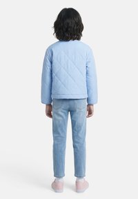 Παιδί με μακριά μέχρι τους ώμους σκουρόχρωμα μαλλιά, φορούσε γαλάζιο πάλλευκο μπουφάν με καπιτονέ ύφασμα, γαλάζια τζιν παντελόνια, άσπρες κάλτσες και ροζ αθλητικά παπούτσια, κοιτάζει προς τα πίσω.