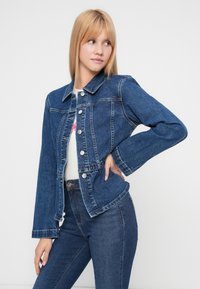 Τζιν μπουφάν σε σκούρο μπλε με κουμπιά κλεισίματος, μυτερό γιακά και ελαφρώς φαρδιές μανσέτες. Διαθέτει δύο μπροστινές τσέπες και εφαρμοστή σχεδίαση.